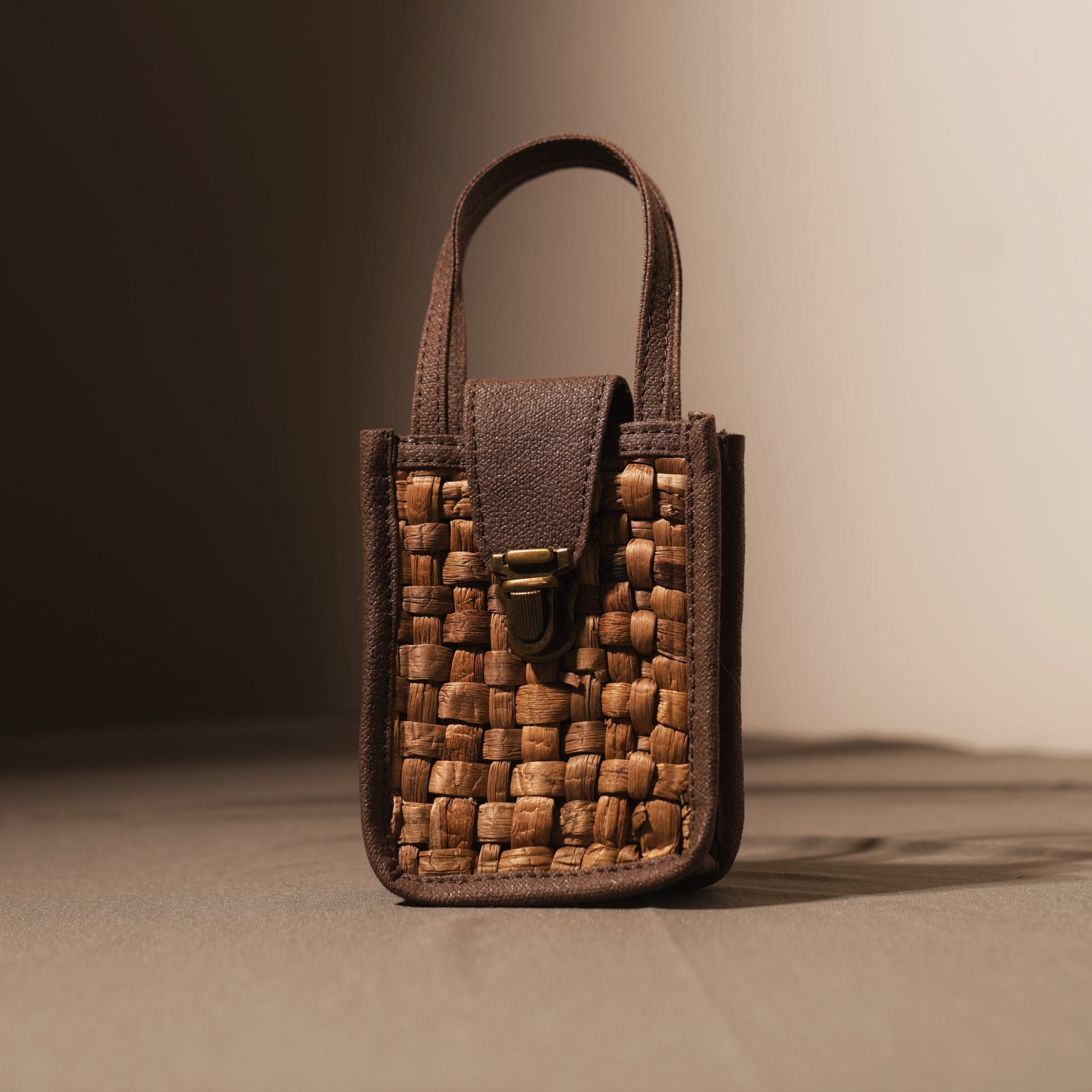 
A close-up shot of a small, rectangular bag made from woven natural fibers in a light brown hue, framed by darker brown leather. The bag has a top handle and a flap closure secured by a metallic clasp. The woven texture of the bag is prominent, showcasing individual strands.