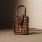 
A close-up shot of a small, rectangular bag made from woven natural fibers in a light brown hue, framed by darker brown leather. The bag has a top handle and a flap closure secured by a metallic clasp. The woven texture of the bag is prominent, showcasing individual strands.